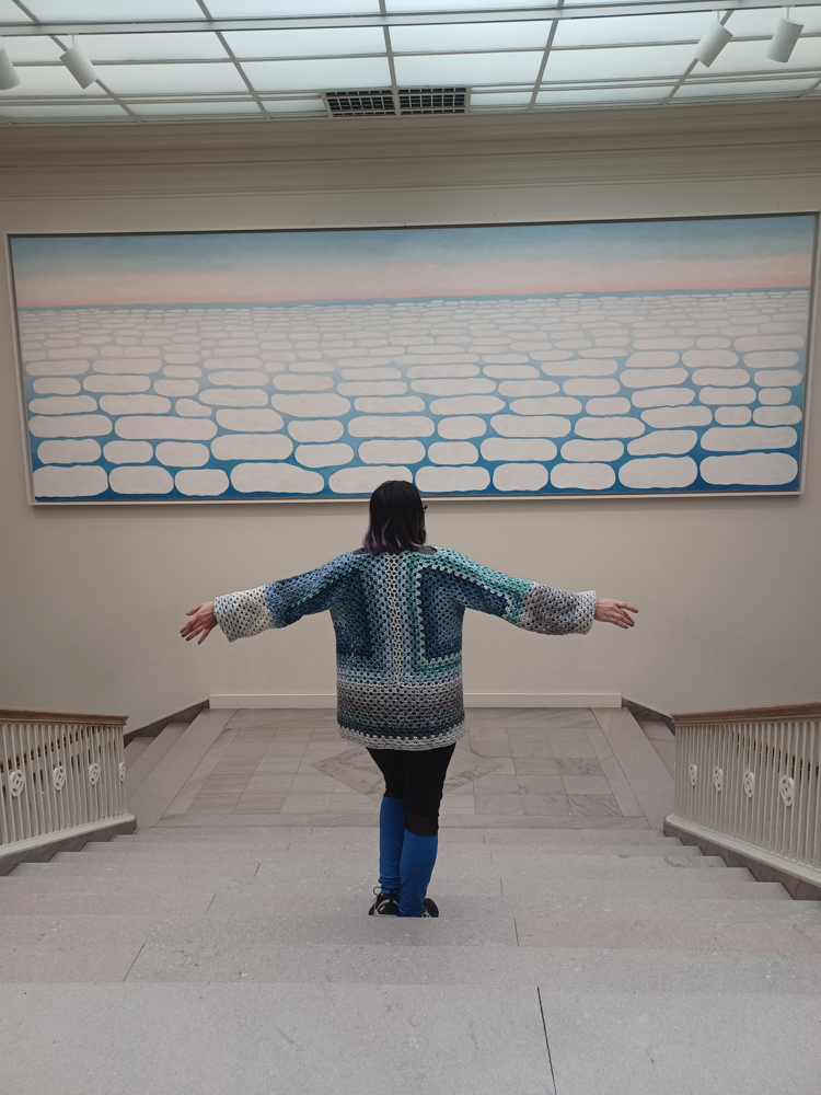 Abigail in a gallery, arms outstretched before a large cloud painting, wearing a crocheted cardigan that echoes it